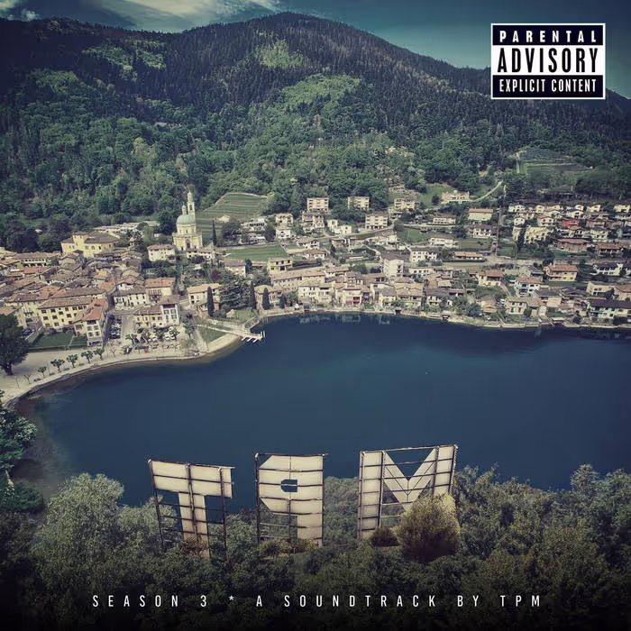Aerial view of a lakeside town with green forested hills, showing a reversed 'TPM' sign among trees and a parental advisory label in the top right corner.